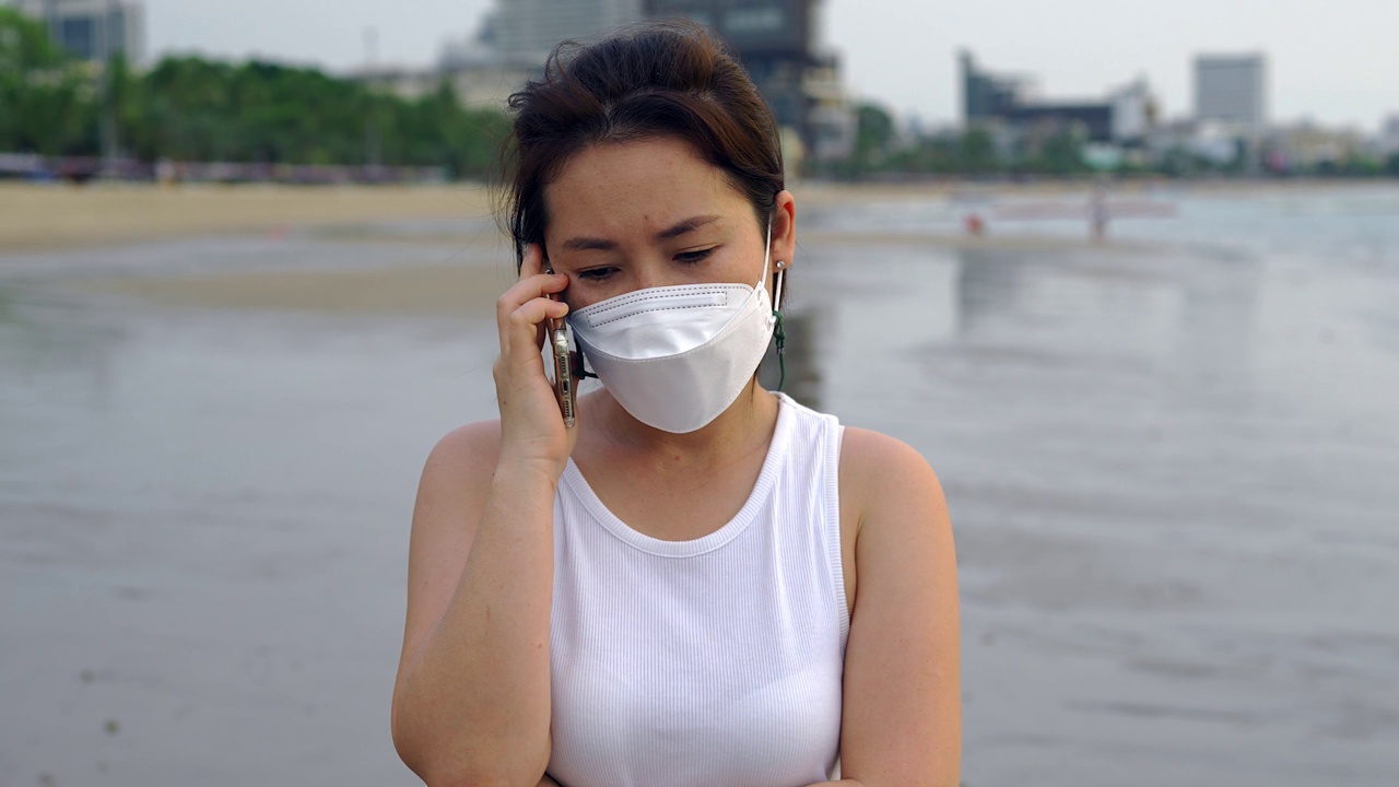 沙滩上，一名年轻女子戴着防护口罩，面带表情地打电话。视频素材