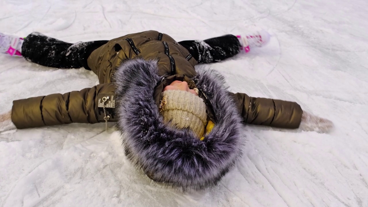 小女孩穿着冰鞋躺在冰上做雪天使。喜庆的心情，圣诞节、新年、节假日，活跃的冬季运动。慢动作视频素材