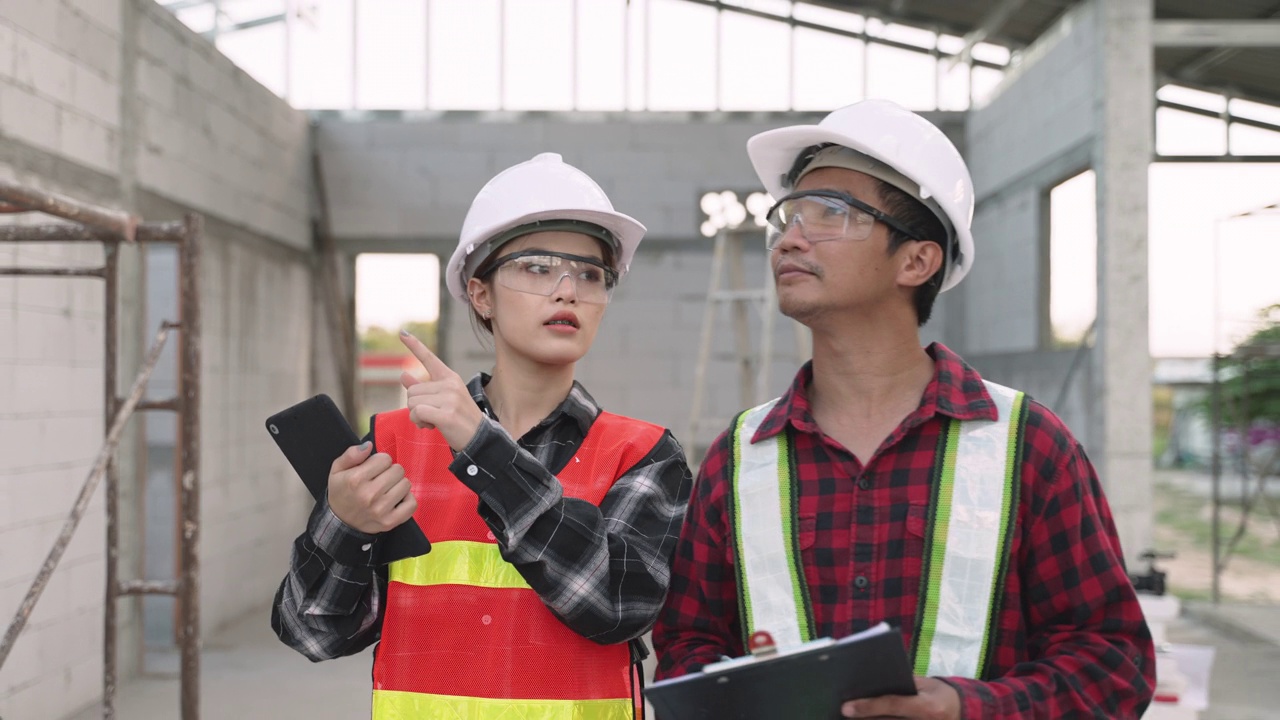 亚裔女建筑师和男承包商在施工现场，戴安全帽和护目镜，手持写字板和剪贴板行走，进行建筑勘察。视频素材