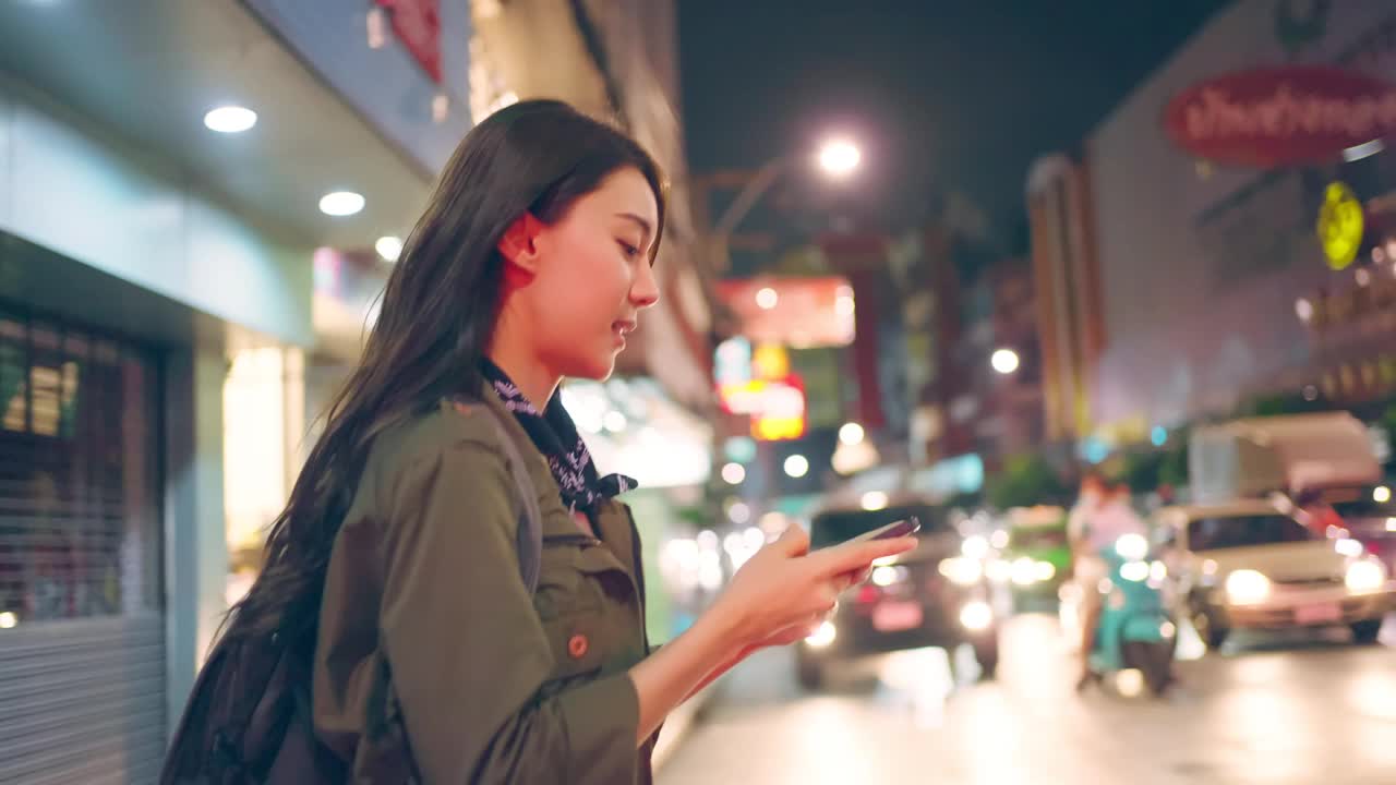 亚洲美丽的女人背包客滚动手机，而走在城市。有吸引力的女孩旅游旅行者独自在街上旅行，使用智能手机享受假期假期旅行的时间在夜间道路视频下载