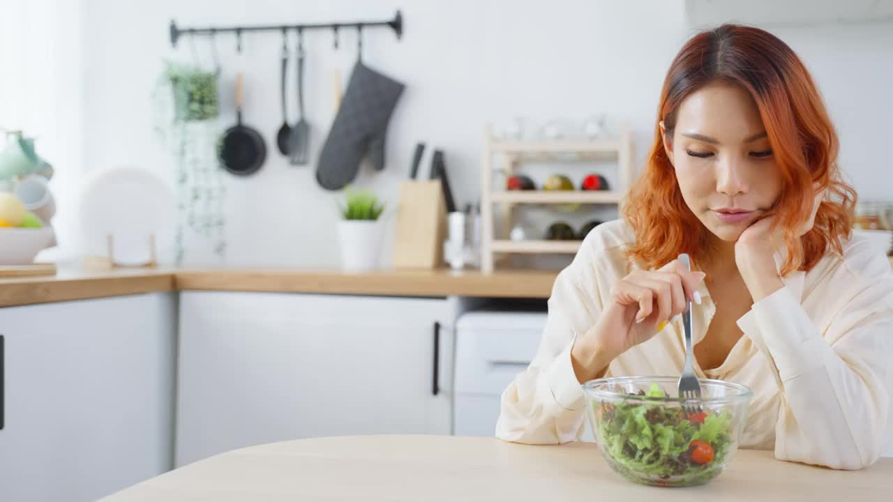 亚洲有吸引力的女人在家里厨房的桌子上吃绿色沙拉。年轻美丽的素食女孩感到快乐，享受早餐蔬菜健康的食物，在家里减肥健康。视频素材