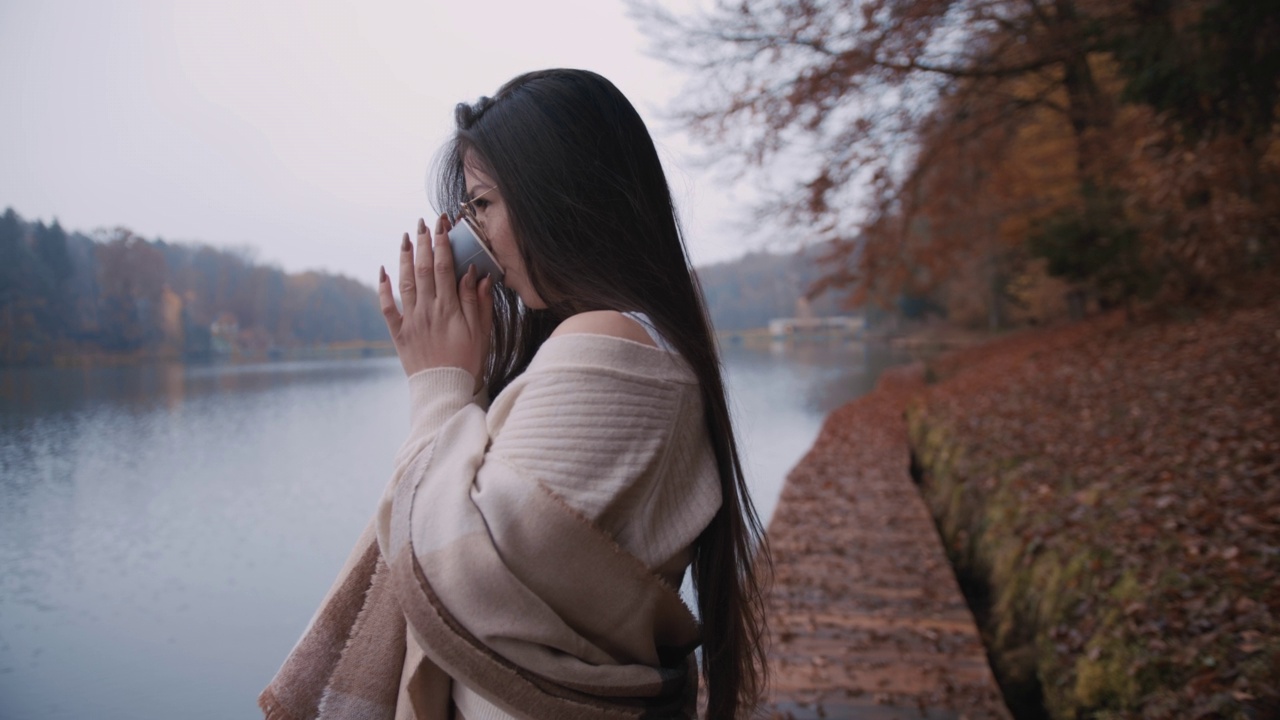 一名年轻女子站在湖边的木板路上，用纸杯喝着热咖啡视频素材