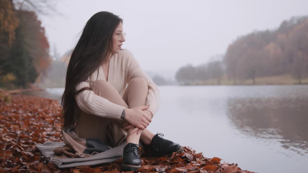 年轻女子坐在湖边的木板路上，上面铺着枯叶视频素材