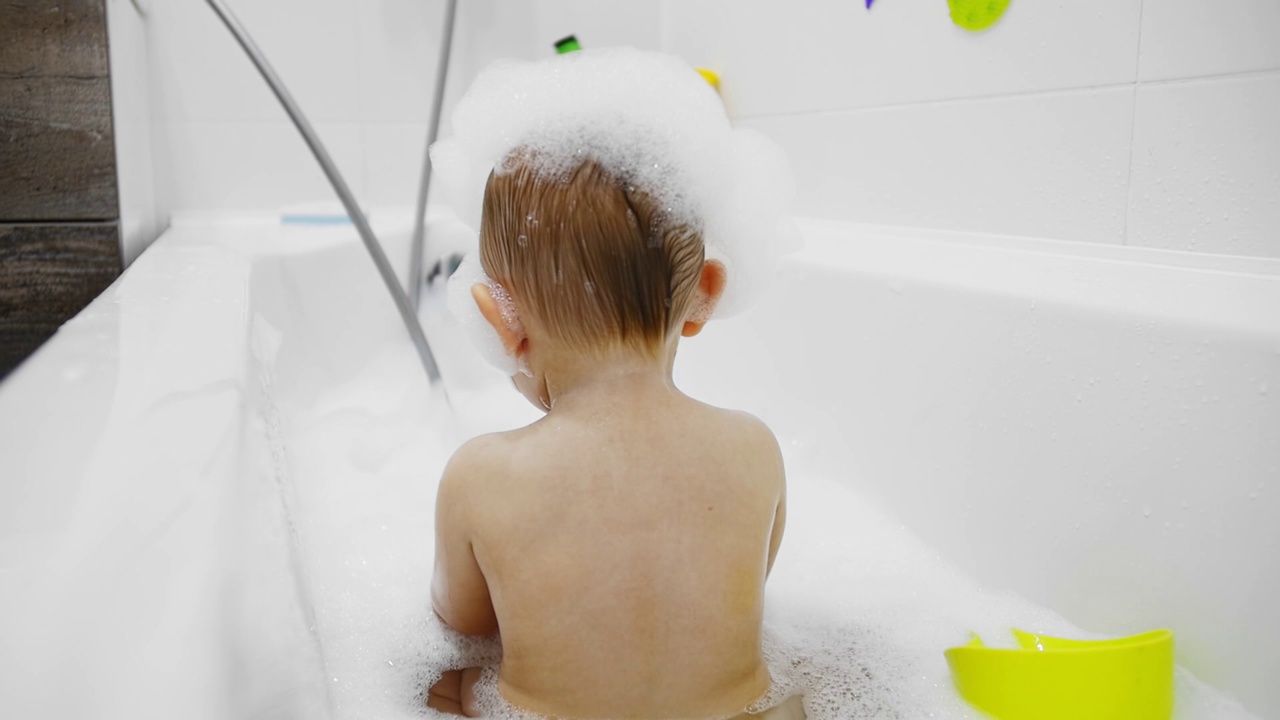 一个胖乎乎的孩子头上沾着泡沫，在白色的浴缸里洗澡视频素材