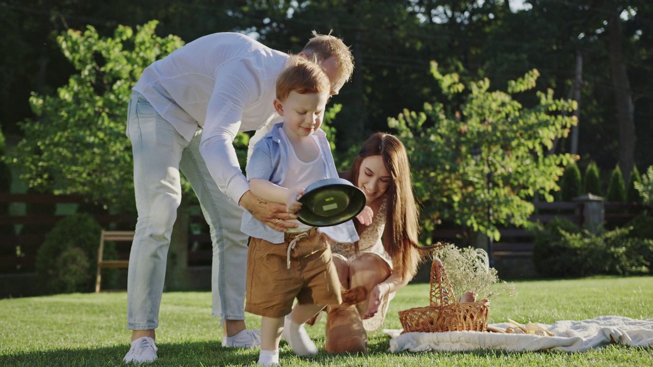 家庭野餐坐在绿色的草地上，在院子里玩一只小狗快乐的笑着儿子走在他的手中一个碗视频下载