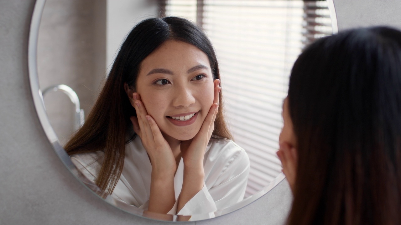 护肤概念。美丽年轻的亚洲女性看着镜子和触摸脸视频购买