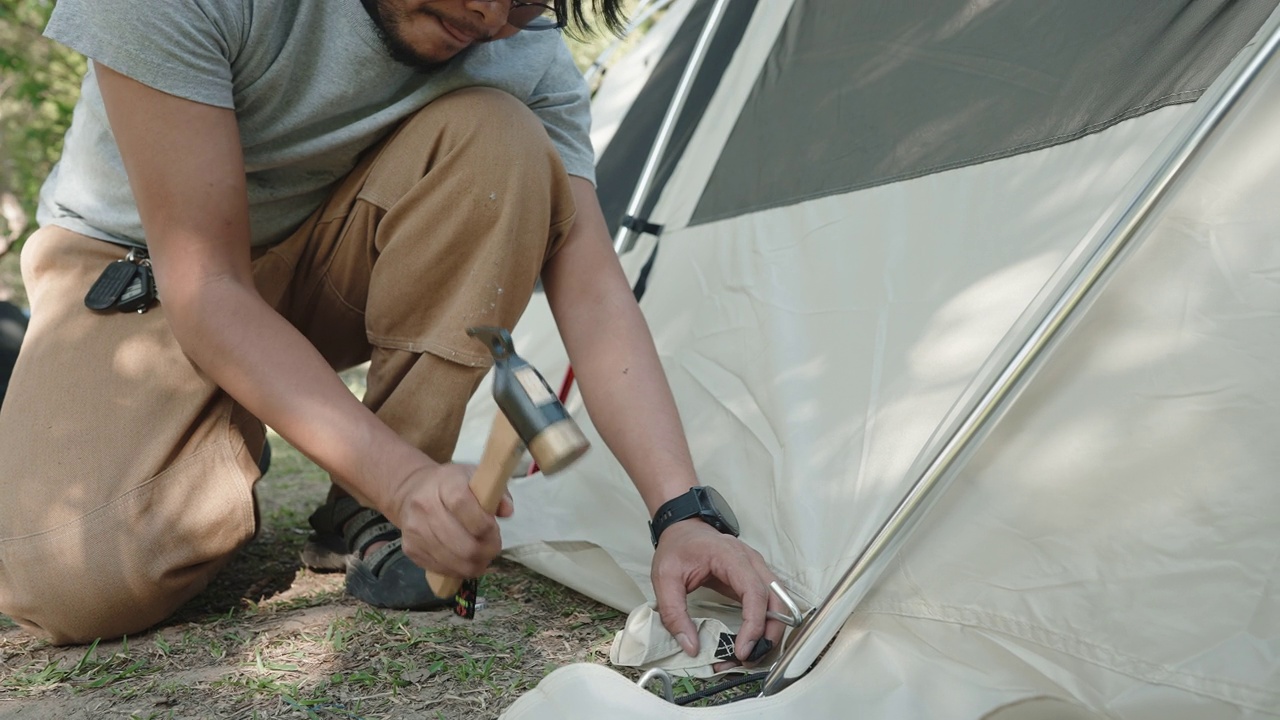 亚洲青年成人露营者用锤子砸露营帐篷的钉子，把帐篷撑在地上视频素材