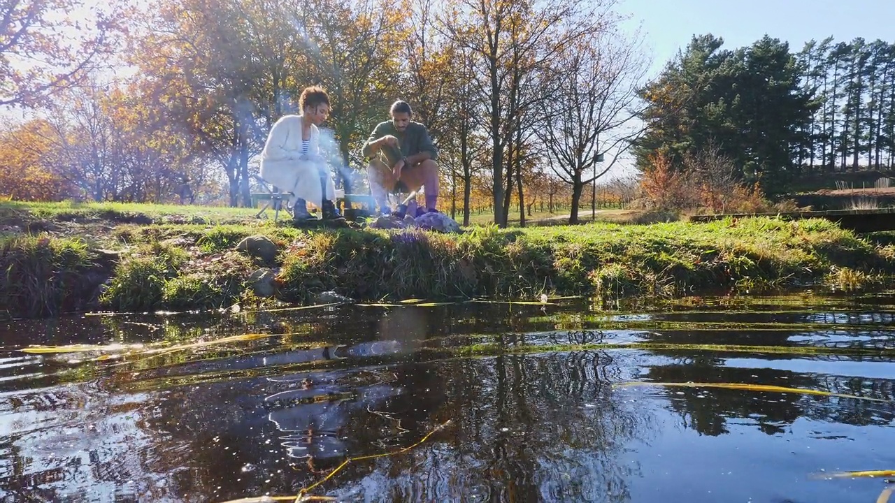 在阳光明媚的秋日湖畔露营地，一对年轻夫妇在篝火旁放松视频素材