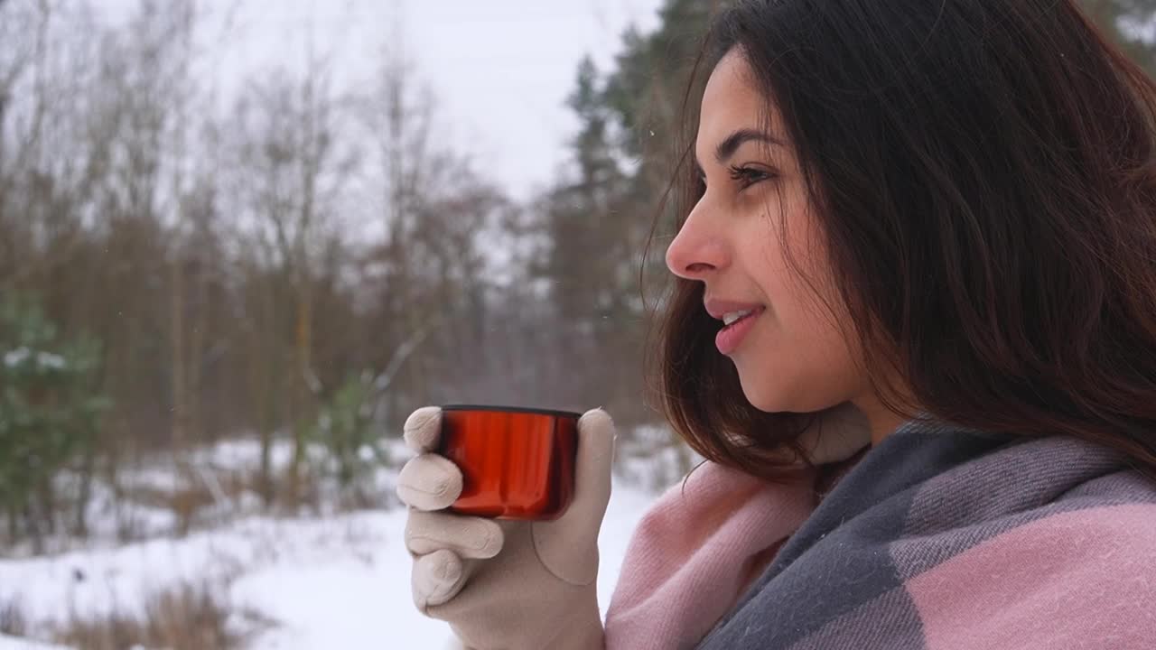 年轻可爱的黝黑黑发女人喝热饮料从橙色保温杯在雪天的冬季森林视频素材