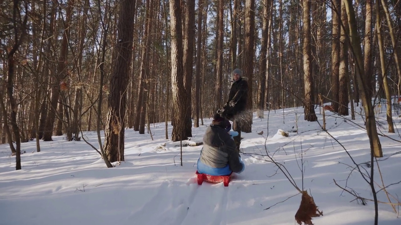 快乐的爸爸带着儿子坐在红色的雪橇上，在阳光明媚的雪原森林里。孩子把雪。视频素材