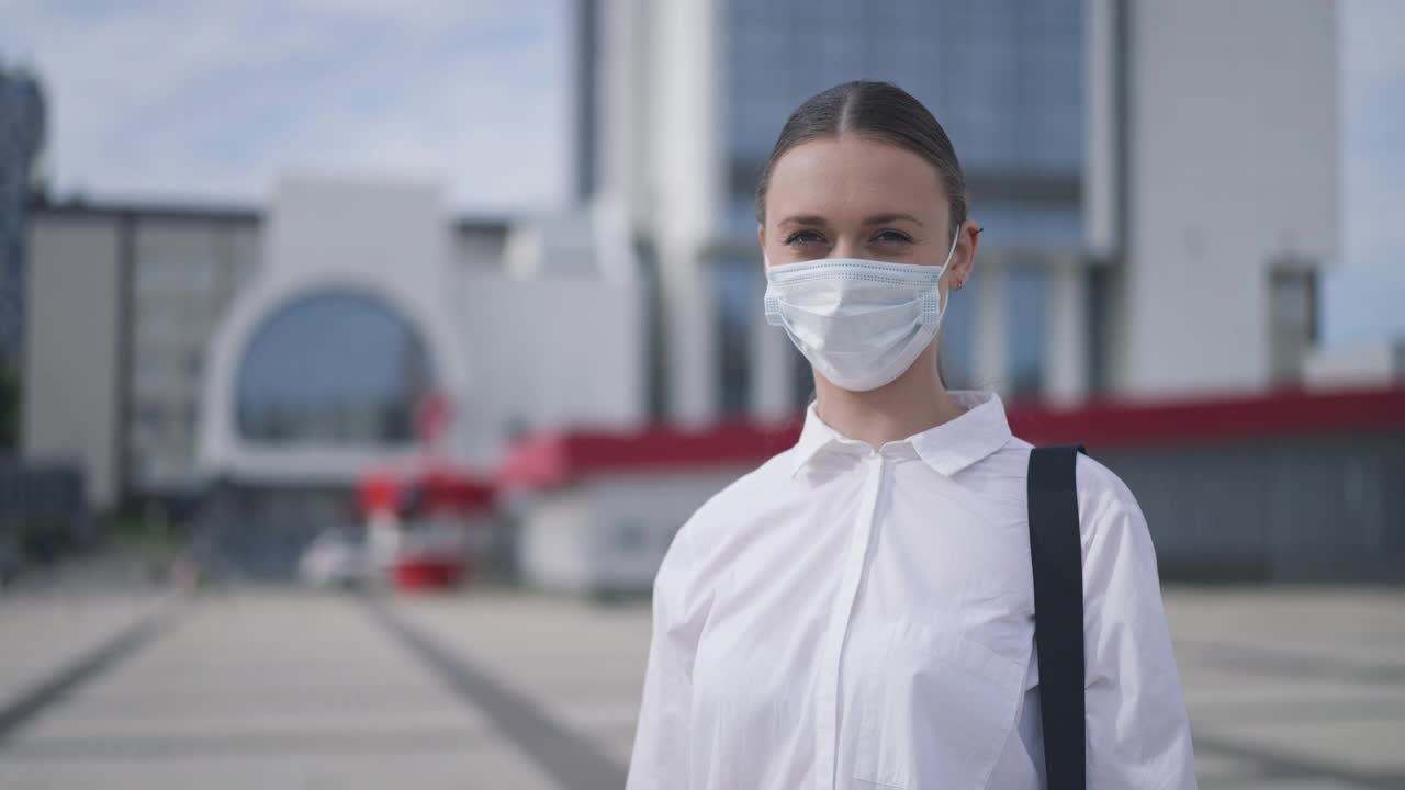 戴着冠状病毒口罩，严肃自信的年轻女子站在城市广场上，双手交叉，没有任何手势。在Covid-19大流行期间，白人苗条女士在市中心摆姿势的慢镜头。视频素材