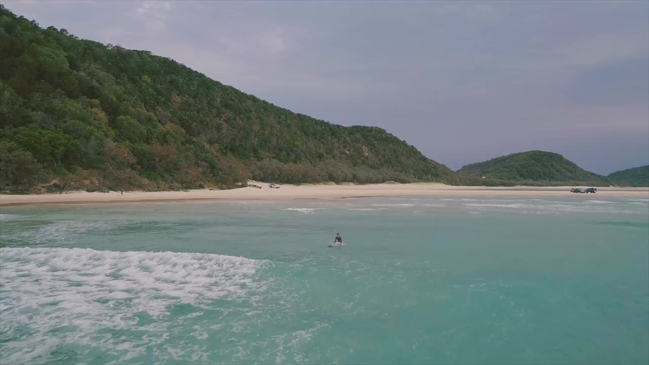 一个美丽的空中/无人机拍摄的冲浪者在一个孤立的原始热带海滩天堂冲浪，清澈的绿松石水。拍摄于仲夏时节，澳大利亚昆士兰州弗雷泽岛海岸的双岛点视频素材