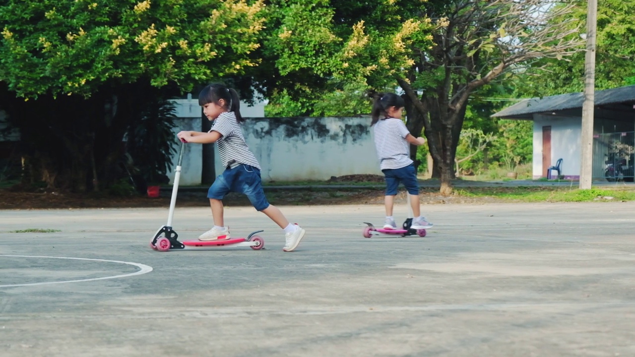 两个可爱的小女孩骑着踏板车在公园户外的夏日道路上。孩子们在户外玩滑板车。儿童积极的休闲和户外运动视频素材