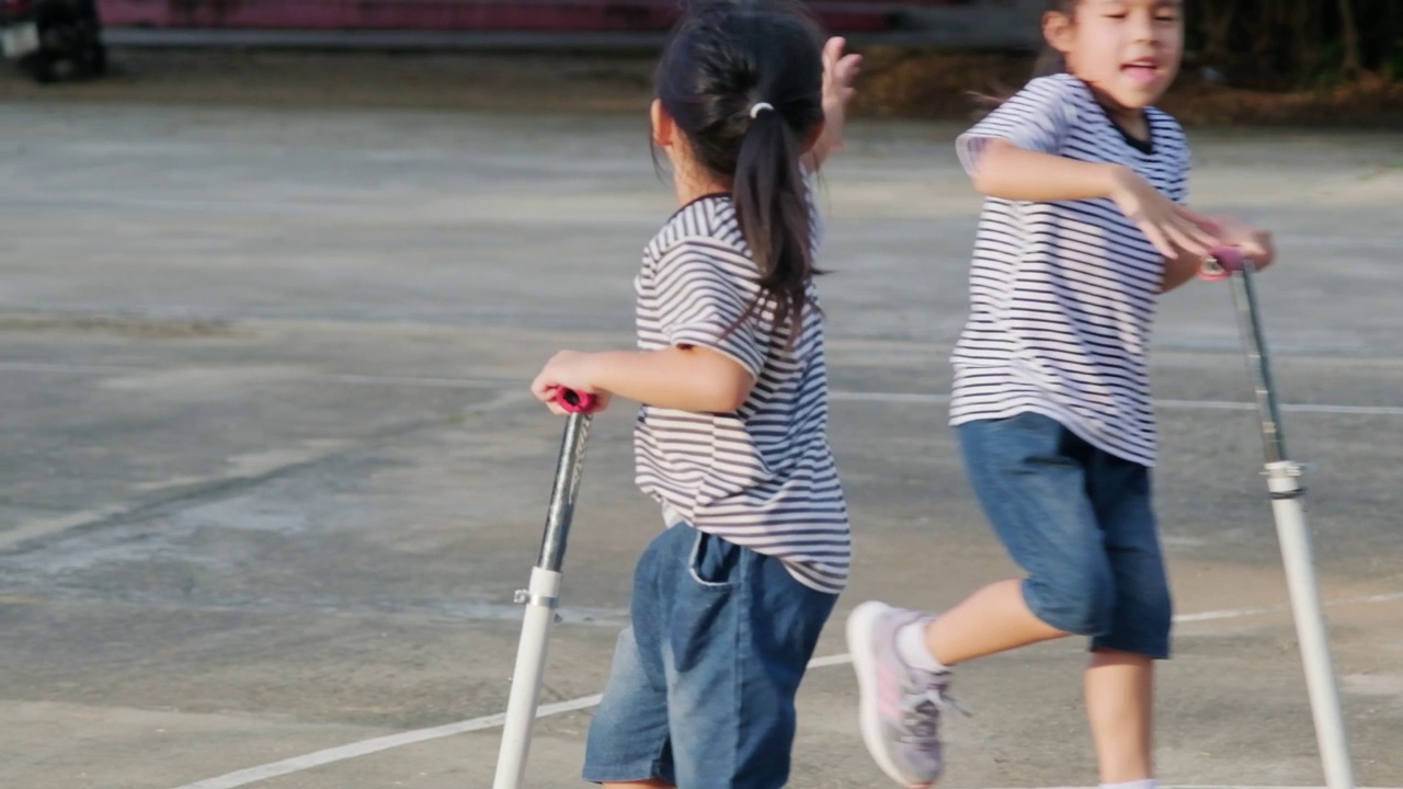 两个可爱的小女孩骑着踏板车在公园户外的夏日道路上。孩子们在户外玩滑板车。儿童积极的休闲和户外运动视频素材