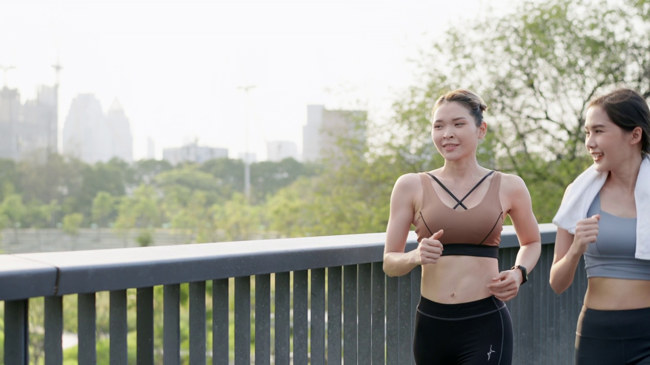 前视图:两个亚洲美丽的中年女性朋友穿着运动服，完美的肌肉一起在公园花园的人行天桥上积极慢跑，靠近中心城市，在早上，积极的生活方式视频素材