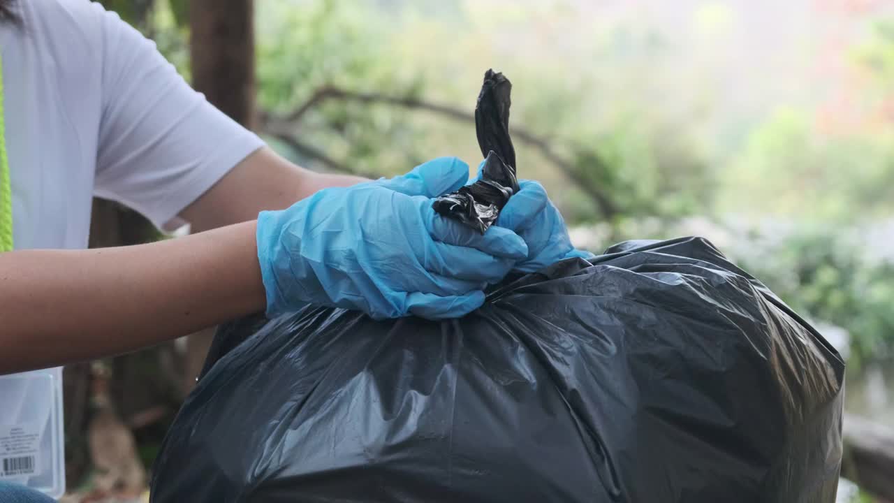 女志愿者在河边捡垃圾后，用手捆住垃圾袋。把垃圾放在垃圾箱里，帮助保护环境。世界环境日视频素材