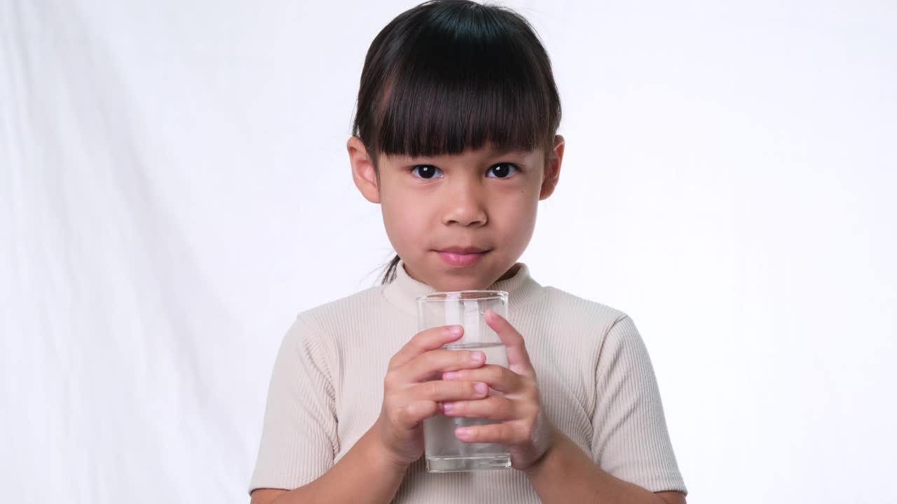 可爱的亚洲小女孩从杯子里喝水，在工作室的白色背景上竖起大拇指。儿童良好的健康习惯。医疗保健的概念视频素材