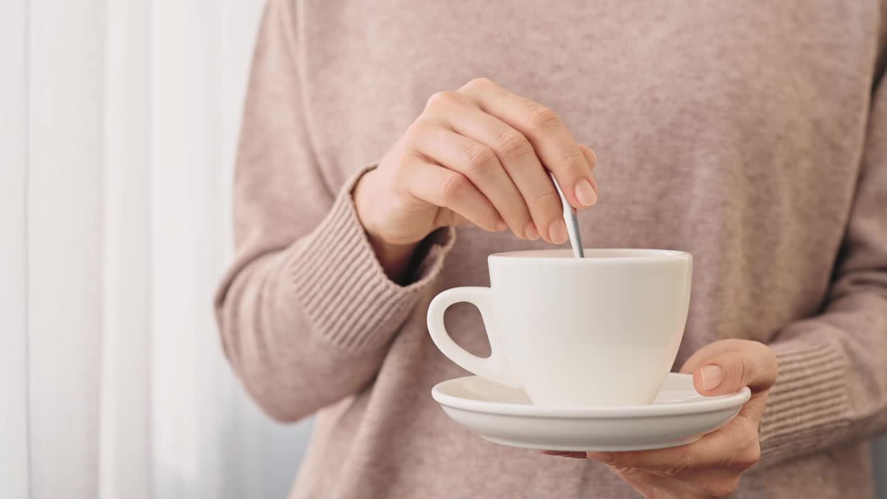 穿着舒适的米色毛衣的年轻女子的手搅拌着一杯茶或咖啡的勺子靠近窗户。手与温柔的指甲握杯与茶碟特写视频素材