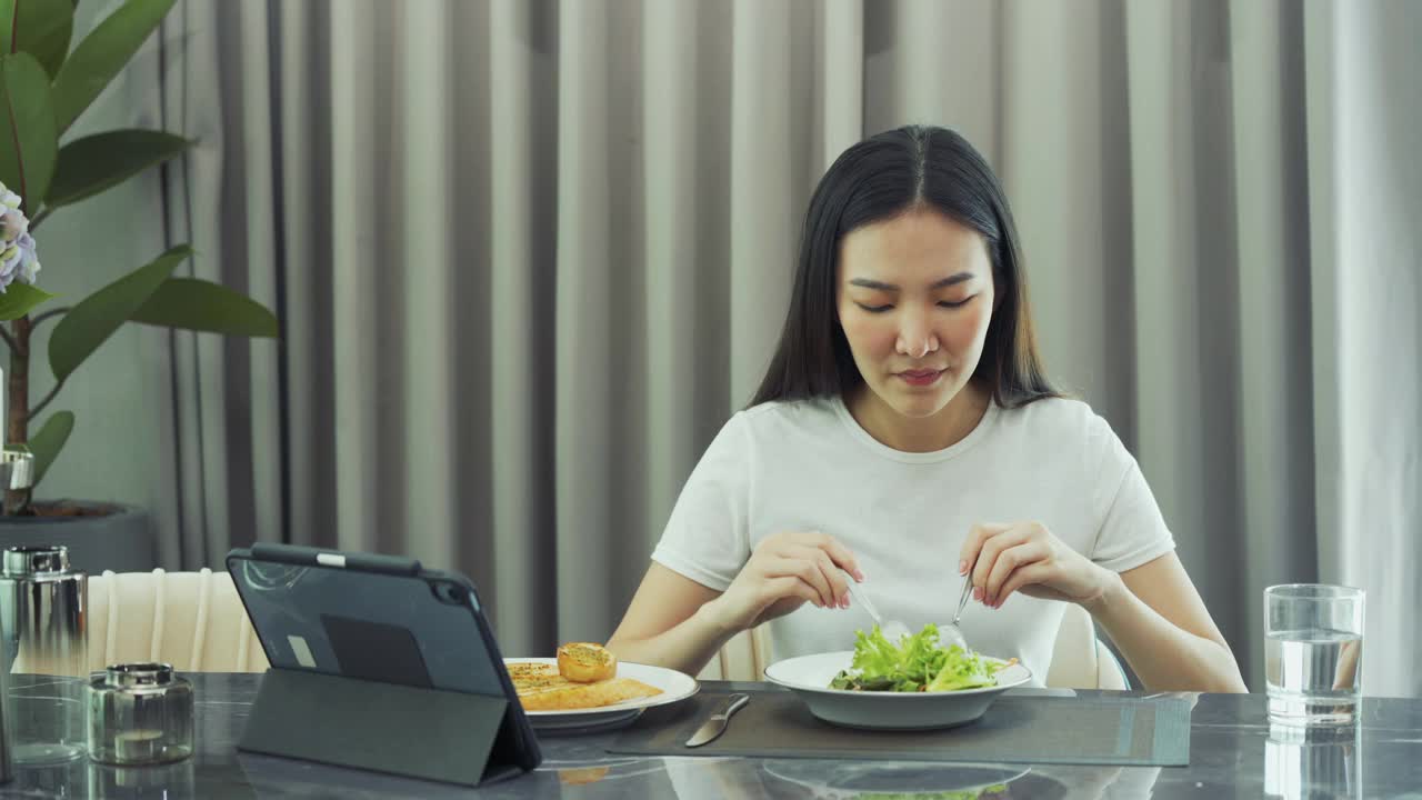 一位亚洲年轻女士在家的时候，一边在平板电脑上拿起她最喜欢的电视剧，一边高兴地把沙拉放进嘴里。视频素材