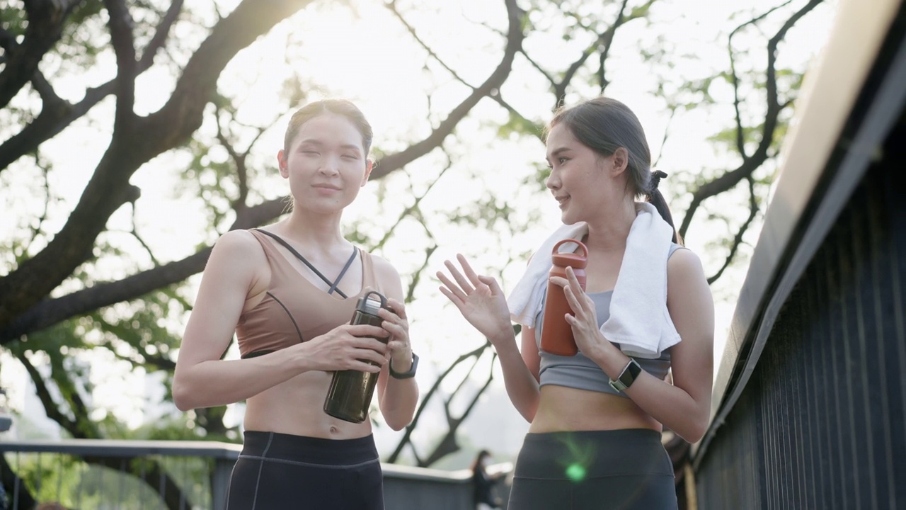 两个亚洲漂亮的中年女性朋友穿着运动服，有着完美的肌肉，在慢跑后休息，从可重复使用的瓶子喝水和交谈，在公园花园的人行桥上，在中心城市附近，积极的生活方式视频素材