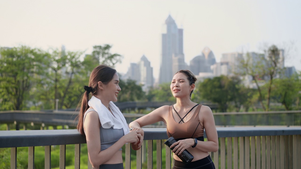 两个亚洲漂亮的中年女性朋友穿着运动服，有着完美的肌肉，在慢跑后休息，从可重复使用的瓶子喝水和交谈，在公园花园的人行桥上，在中心城市附近，积极的生活方式视频素材