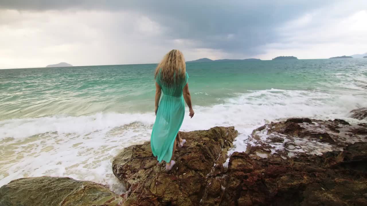 女人走在礁石上的海礁石，暴风雨多云的海洋。蓝色泳衣连衣裙束腰外衣。概念休息，热带度假海岸线旅游夏季假期视频素材