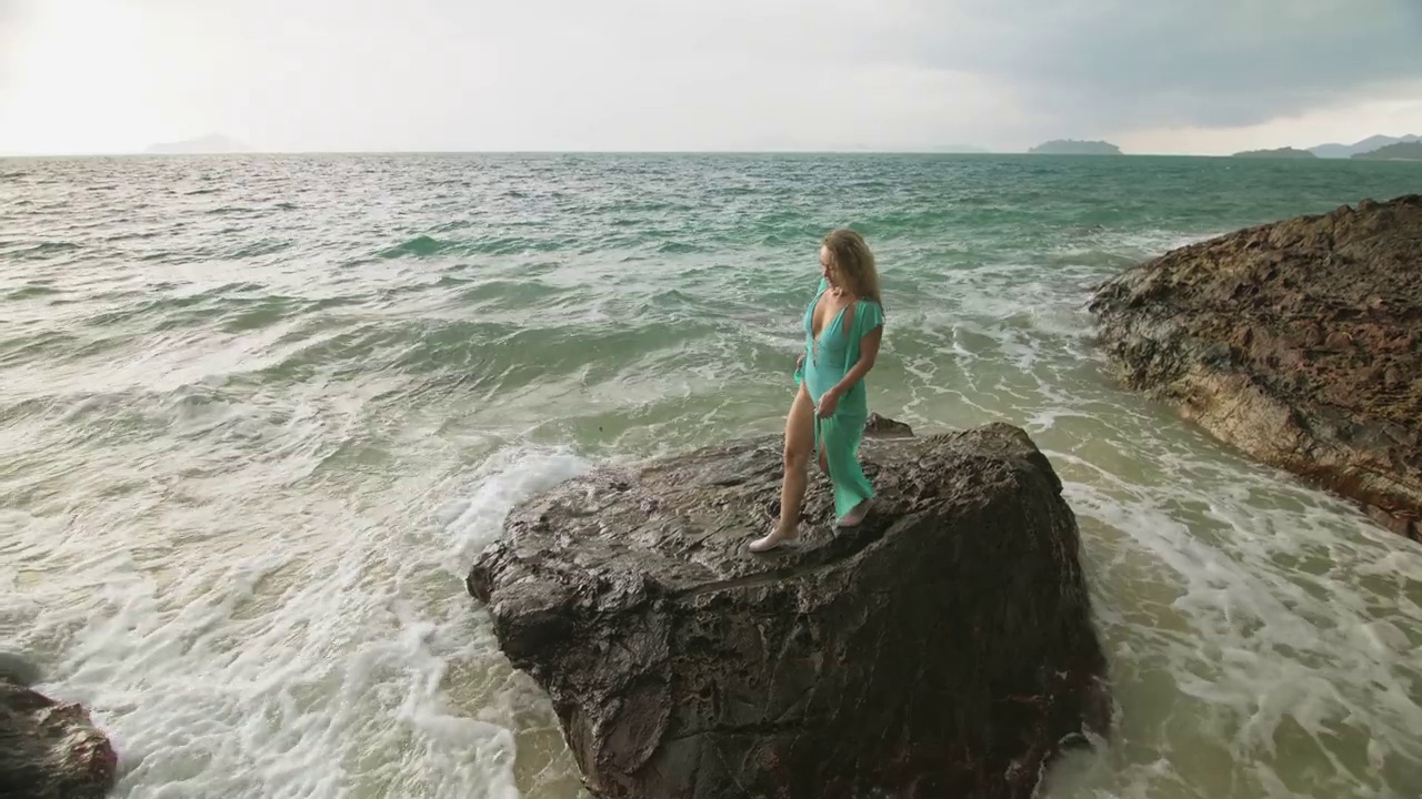 女人走在礁石上的海礁石，暴风雨多云的海洋。蓝色泳衣连衣裙束腰外衣。概念休息，热带度假海岸线旅游夏季假期视频素材