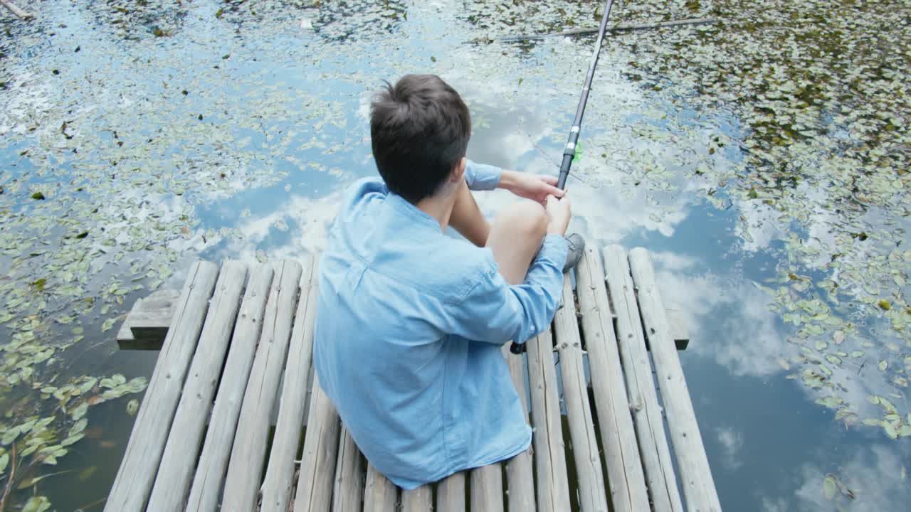 夏天，一个少年拿着鱼竿坐在码头上钓鱼视频素材