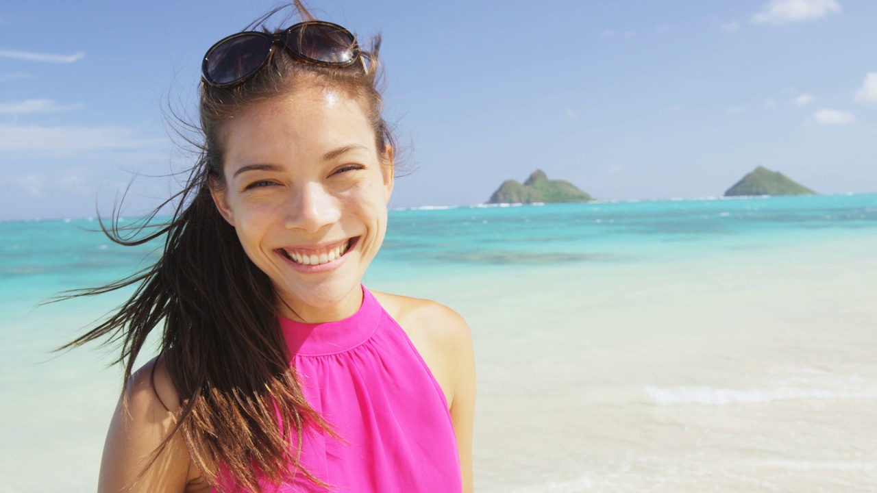 沙滩上的女人肖像。夏威夷拉尼凯海滩的多种族模特。年轻的混血儿亚裔华人/白种人女性穿着粉红色的太阳裙，微笑着看着镜头在夏季旅行假期视频素材