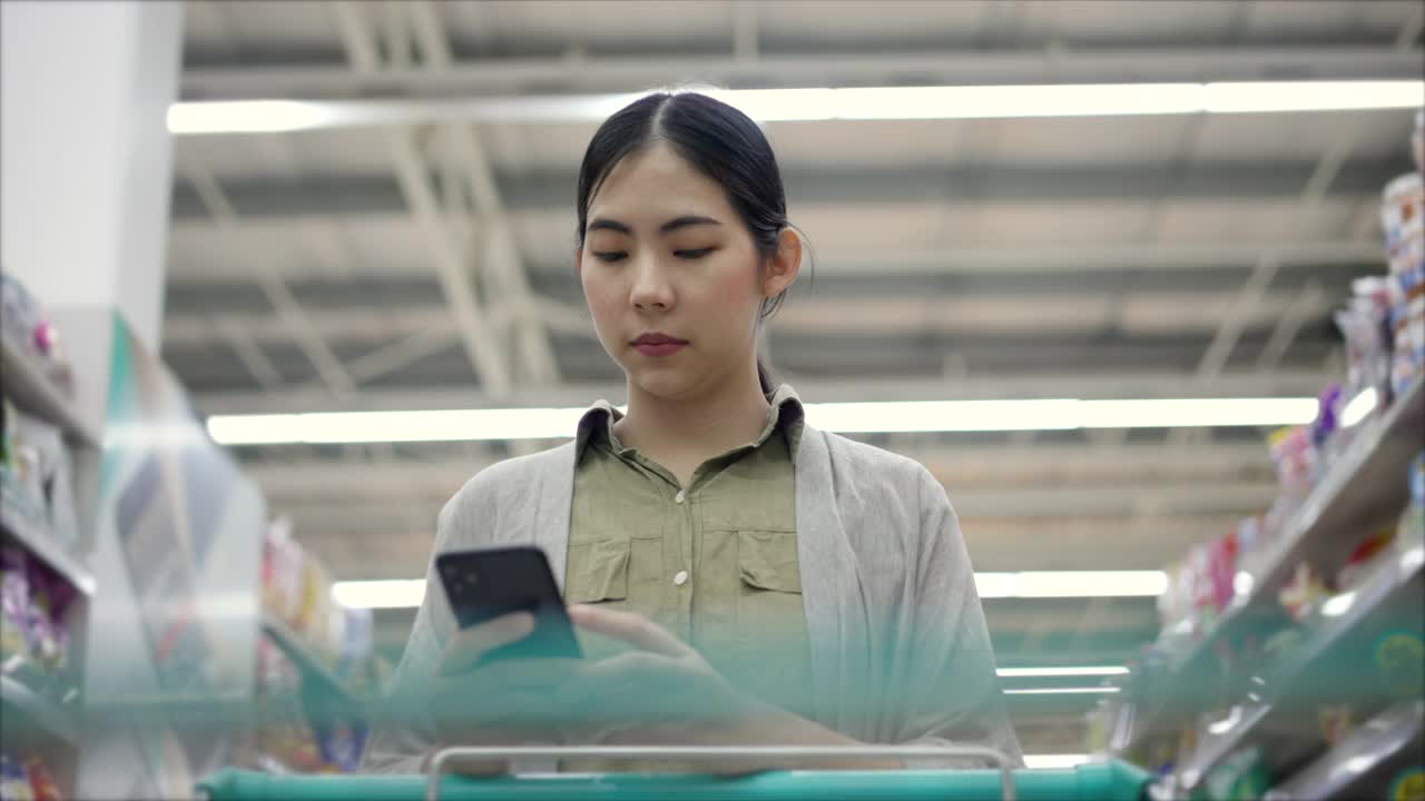 亚洲妇女在超市用智能手机查看购物清单。女性使用智能手机和应用程序在商店检查产品信息。视频素材