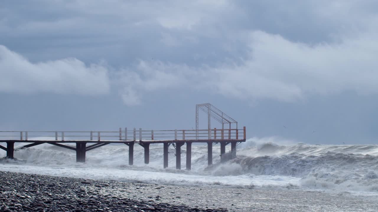 巨大的海浪撞击着海岸和码头蓝色的水背景。海面上的风暴和汹涌的海浪溅起泡沫。刮风坏天气慢动作视频素材