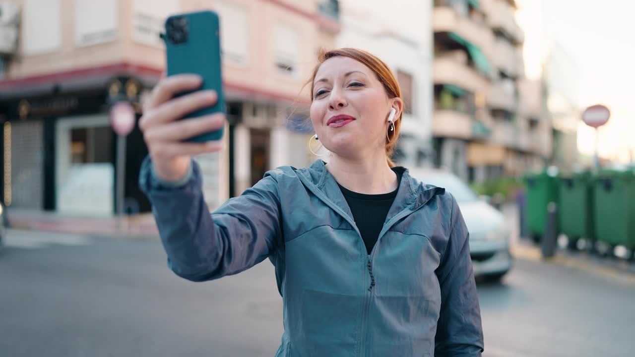 年轻的红发女人穿着运动服在街上打视频电话视频素材