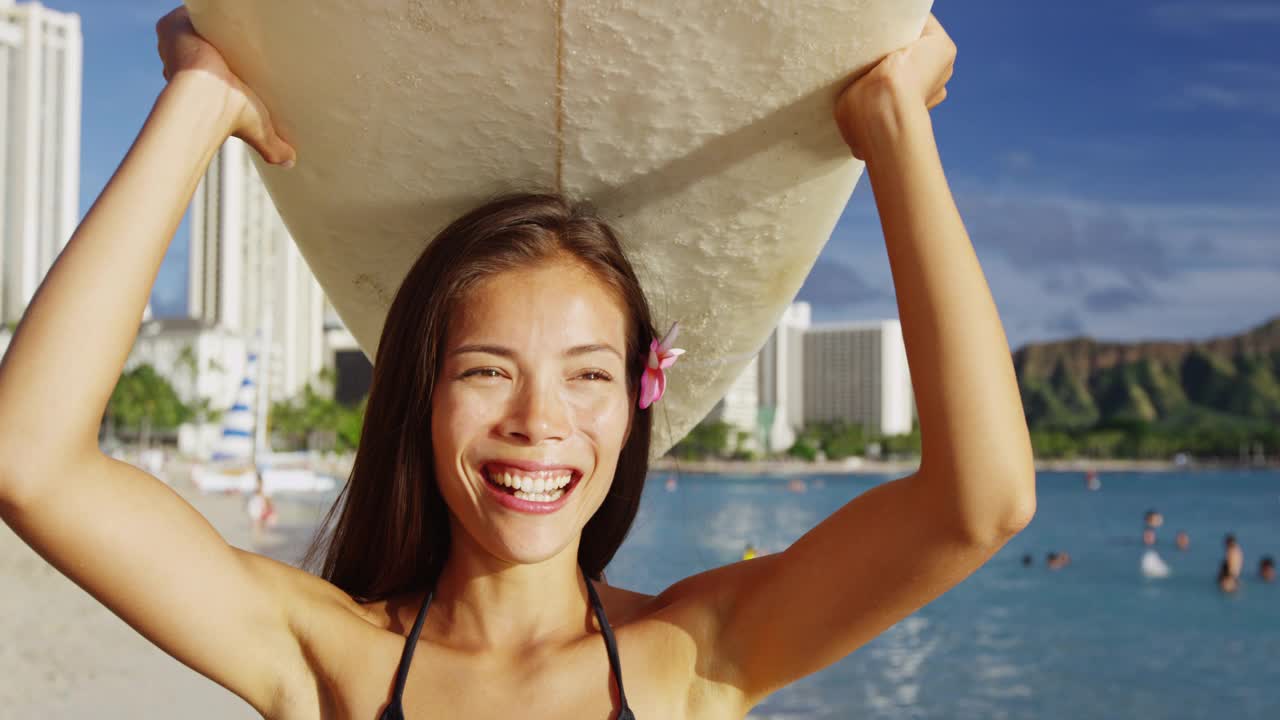 在威基基海滩，一个快乐的年轻女子头顶着冲浪板。暑假期间，美丽的女游客在檀香山的一个小岛上散步。她在天空的映衬下欣赏大海。视频素材