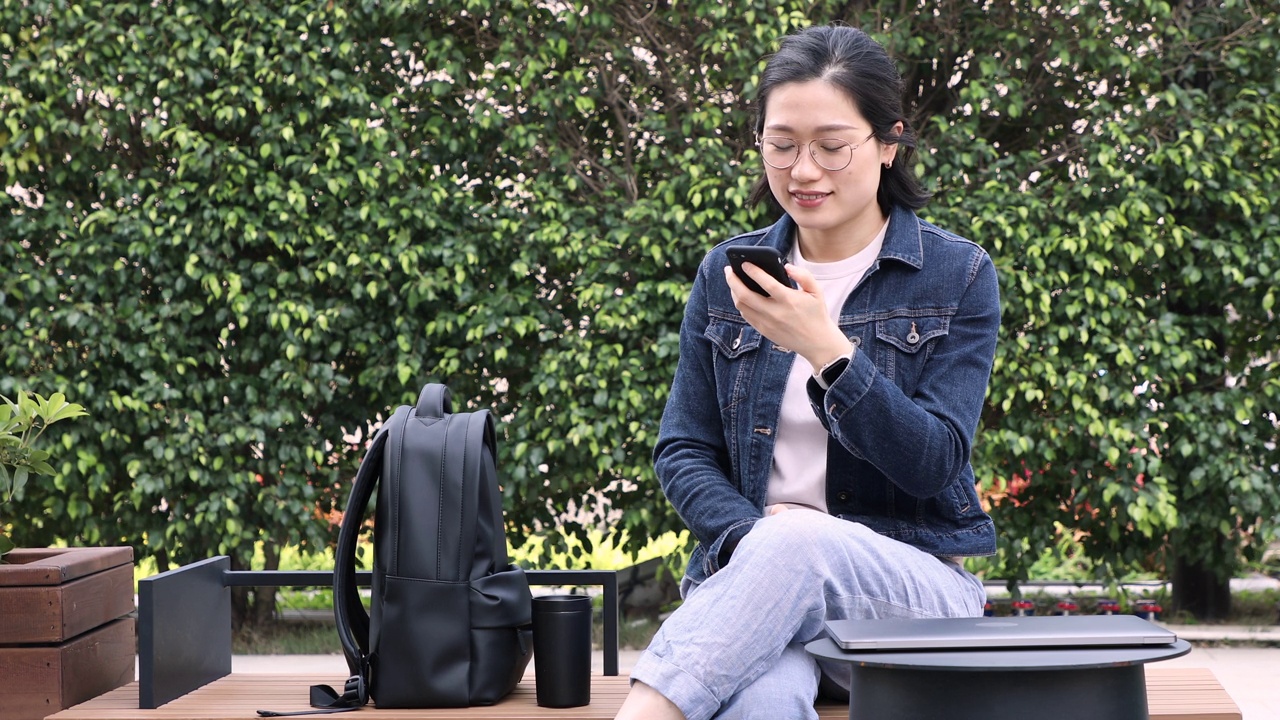 一名亚洲妇女坐在长椅上，一边用可重复使用的杯子喝咖啡，一边打电话视频素材