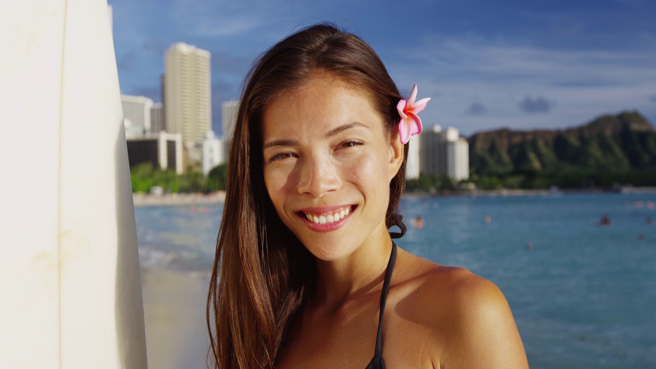 微笑的年轻女子在威基基海滩冲浪。美丽的女游客正在檀香山的一个小岛上度假。在高楼和天空的衬托下，她穿着比基尼。视频素材