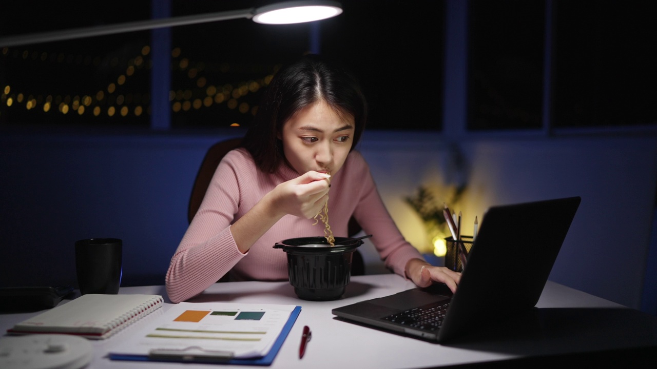 自由亚洲妇女吃面条，晚上在家工作。视频素材