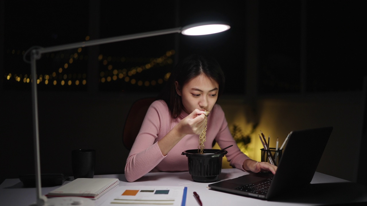 自由亚洲妇女吃面条，晚上在家工作。视频素材