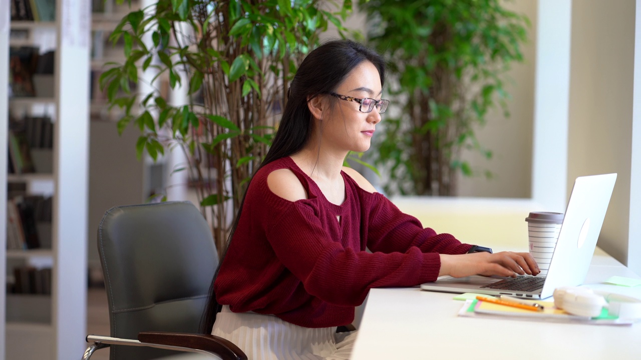 快乐的亚洲学生女孩准备大学考试使用笔记本电脑在图书馆做作业视频素材