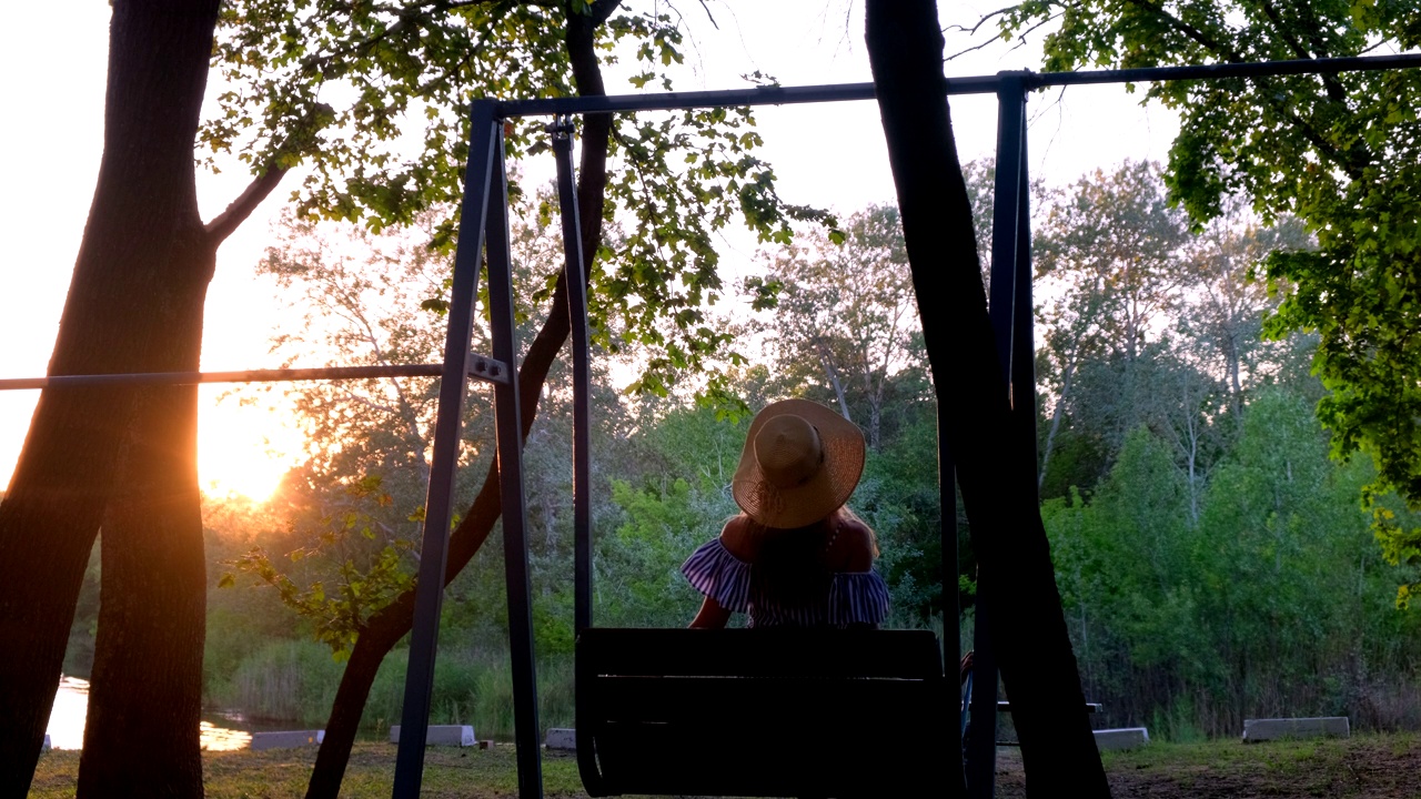 在金色夏日夕阳下荡秋千的女人。快乐的小女孩，戴着草帽，坐在秋千上，看着美丽的大自然视频素材