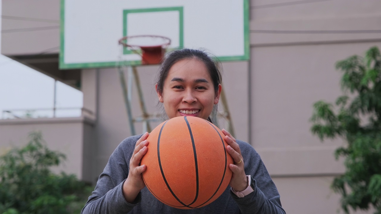 亚洲女子拿着篮球看着镜头在户外篮球场上。视频素材