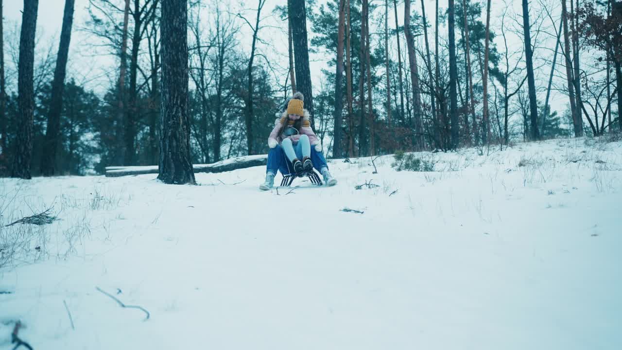 女孩坐在妈妈的腿上，乘着雪橇从雪山上滑下来视频素材