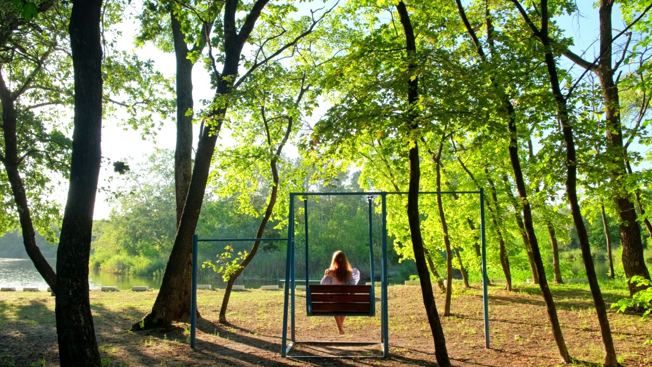 在金色夏日夕阳下荡秋千的女人。快乐的小女孩，戴着草帽，坐在秋千上，看着美丽的大自然视频素材