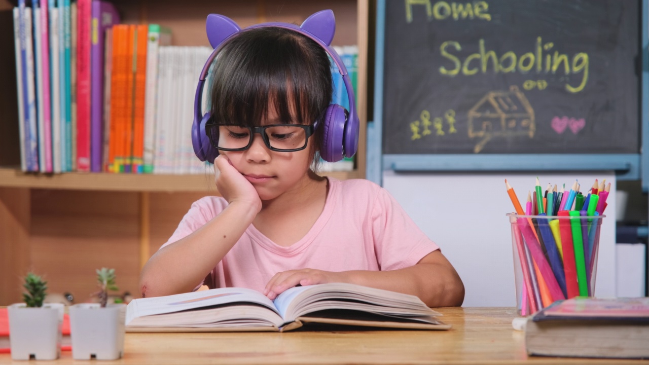 可爱的小女孩戴着耳机听有声书，桌上放着英语学习书。英语学习与现代教育视频素材