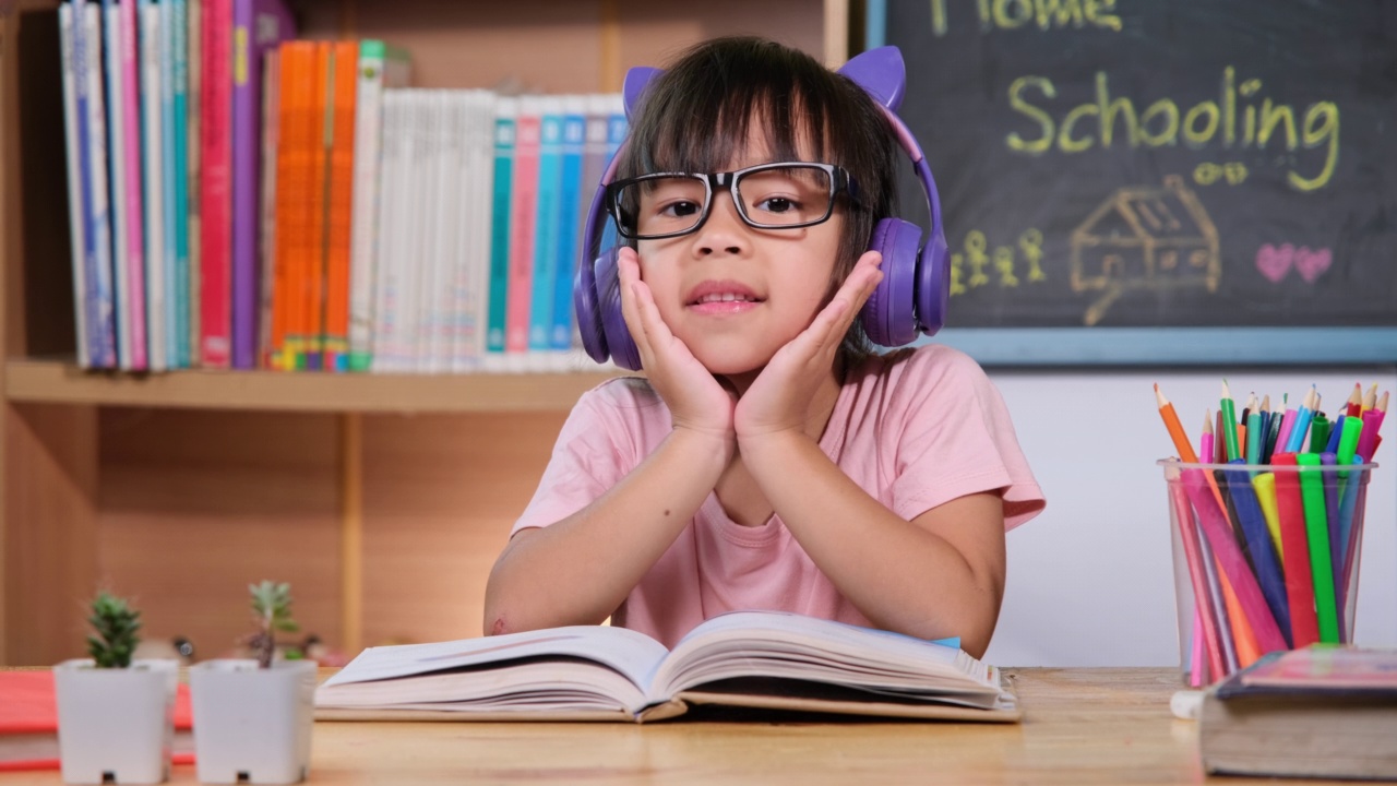 可爱的小女孩戴着耳机听有声书，桌上放着英语学习书。英语学习与现代教育视频素材