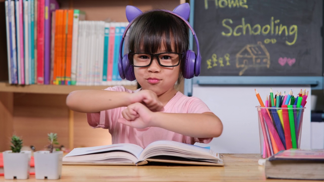 可爱的小女孩戴着耳机听有声书，桌上放着英语学习书。英语学习与现代教育视频素材