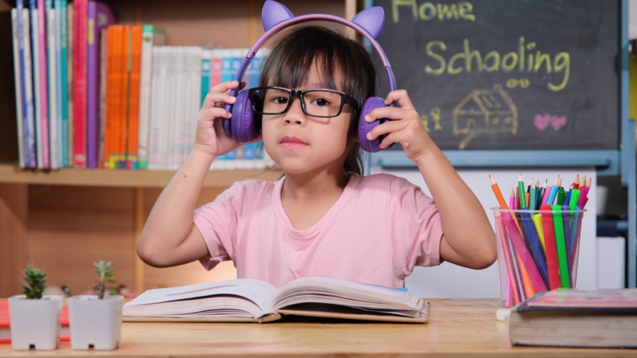 可爱的小女孩戴着耳机听有声书，桌上放着英语学习书。英语学习与现代教育视频素材