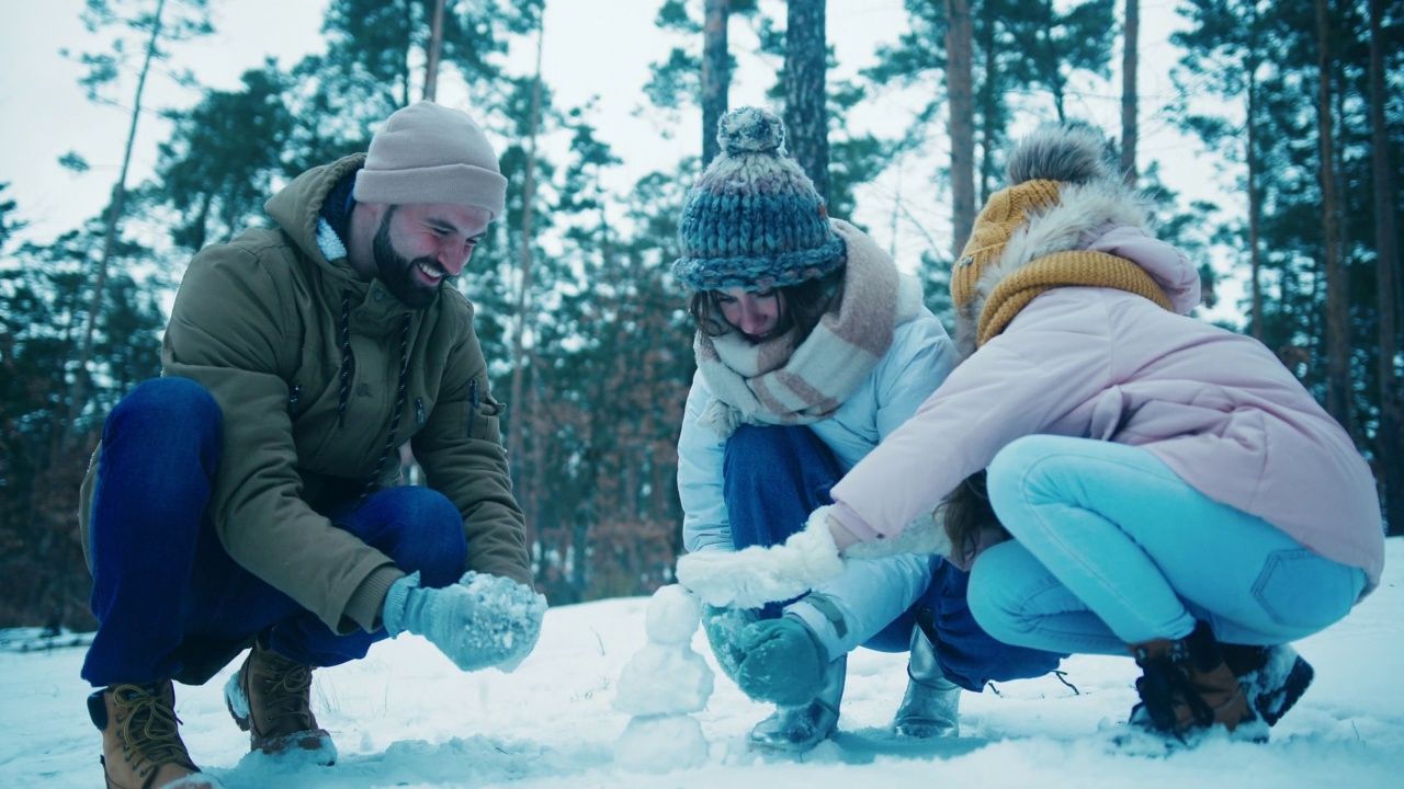 年轻的一家人在松树林空地上堆小雪人视频素材