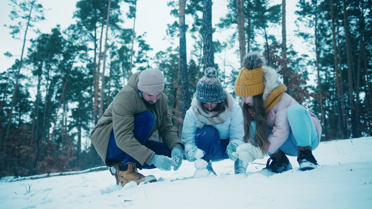 年轻的一家人在松树林空地上堆小雪人视频素材