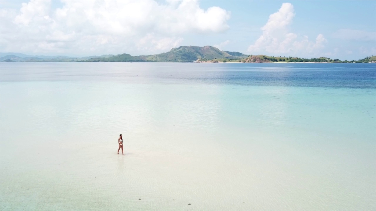 年轻的比基尼女子走在白色沙滩附近的蓝色热带海洋海浪。视频素材