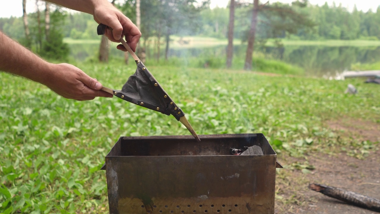 一个人在烧烤时用硬木壁炉风箱生火。在湖边钓鱼的树林里休息视频素材